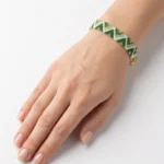 دستبند میوکی سبز بسته شده روی مچ دست با طرح زیگزاگ و دانه‌های طلایی