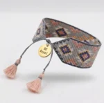 نمای کامل دستبند بافت میوکی طرح لوزی خاکستری و بنفش با آویز مدال طلایی و منگوله‌های نخی صورتی