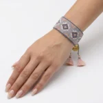 دستبند بافت میوکی طرح لوزی خاکستری و صورتی بسته‌شده روی مچ دست با آویز مدال طلایی و منگوله نخی