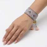 دستبند بافت میوکی طرح لوزی خاکستری و صورتی بسته‌شده روی مچ دست با آویز مدال طلایی و منگوله نخی